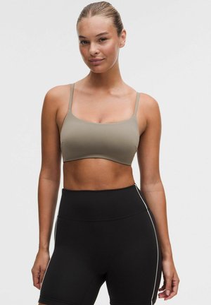 Mujer con un sujetador deportivo beige y pantalones cortos deportivos negros de talle alto frente a un fondo liso.