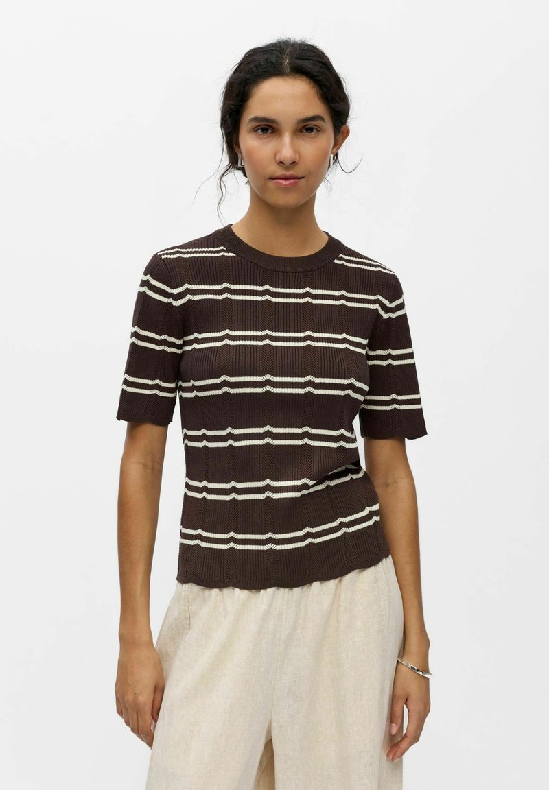 Jeune femme aux cheveux foncés portant un haut tricoté marron à manches courtes avec des rayures blanches horizontales et un pantalon beige clair, debout devant un fond blanc.