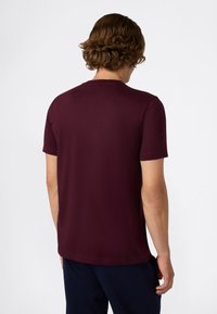 Man met krullend bruin haar in een effen donkerpaars t-shirt met korte mouwen en donkere broek, staand met zijn rug naar de camera tegen een witte achtergrond.