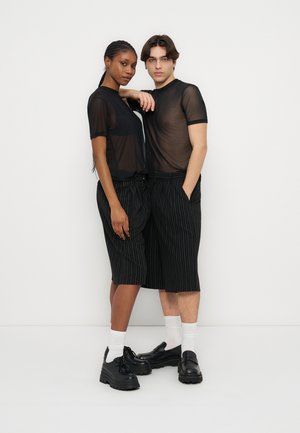 Două persoane care poartă bluze negre transparente, pantaloni scurți negri cu dungi și lungime până la genunchi, șosete albe și pantofi negri masivi, stând în fața unui perete alb simplu.