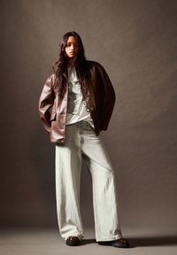 Donna con lunghi capelli scuri che indossa una giacca di pelle marrone, una camicia bianca, jeans larghi chiari e scarpe chunky scure, in piedi contro uno sfondo semplice.