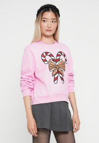 Różowa bluza z graficznym wzorem w postaci cukrowej laski i błyszczącej kokardy. Posiada długie rękawy i ściągacze na mankietach. Połączona z szaro-plisowaną spódnicą.