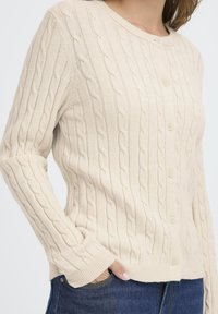 Beige strikket cardigan med kabelmønster, rund halsudskæring, knaplukning og ribbede manchetter, parret med blå denim jeans.