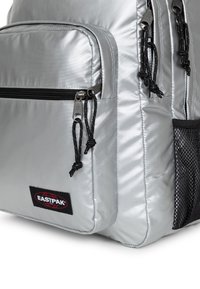 Hopeinen reppu, joka on valmistettu sileästä, heijastavasta materiaalista, jossa on useita vetoketjullisia taskuja, mustat metalliosat ja verkkosivu tasku. Sisältää logopatchin.