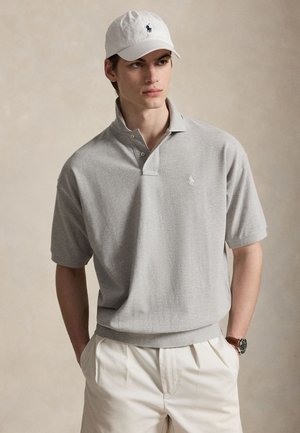 Νέος άνδρας με ανοιχτό γκρι πουκάμισο πόλο, λευκό καπέλο με μαύρο λογότυπο, λευκό παντελόνι και ρολόι χειρός, στέκεται με τα χέρια στις τσέπες.