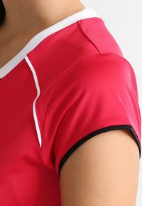 Červené sportovní tričko s krátkým rukávem, s bílými a černými akcenty na límci a okrajích rukávů. Hladká, lesklá textura látky.