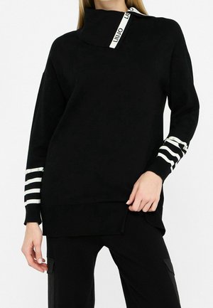 Pull noir à manches longues avec col montant, poignets rayés blancs avec perles, et étiquette de marque sur le col porté avec un pantalon noir.
