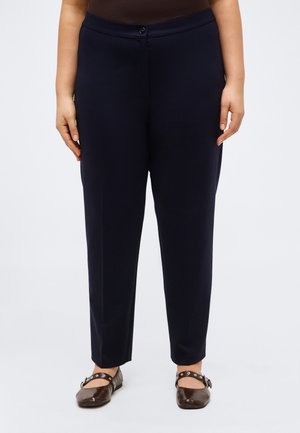 Naine, kes kannab tumesiniseid sirge säärega pükse ja tumepruune Mary Jane tüüpi kingi, seisab ühevärvilise heleda tausta ees.