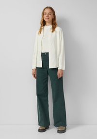 Leichtes Creme-Hemd mit Knöpfen, kombiniert mit dunkelgrünen, weit geschnittenen Hosen und braunen Stiefeletten. Glatte Stoffe mit lässiger Passform.