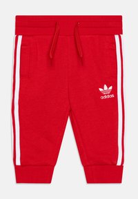 Rdeče trenirke z belimi stranskimi črtami, narejene iz mehke tkanine. Imajo nastavljiv pas z vrvico in stranske žepke. Prikazana je Adidasova oznaka.