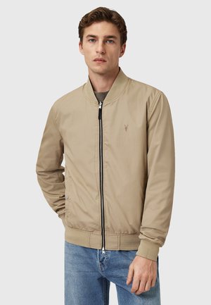 Beige bomberjakke med lynlås foran, ribbede manchetter og krave. Bemærkelsesværdig detalje: lille logo på venstre bryst. Bæres med blå jeans.