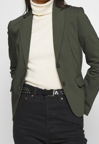Olive green blazer with structured shoulders, two buttons, flap pockets, and a tailored fit, paired with a white sweater and black jeans.