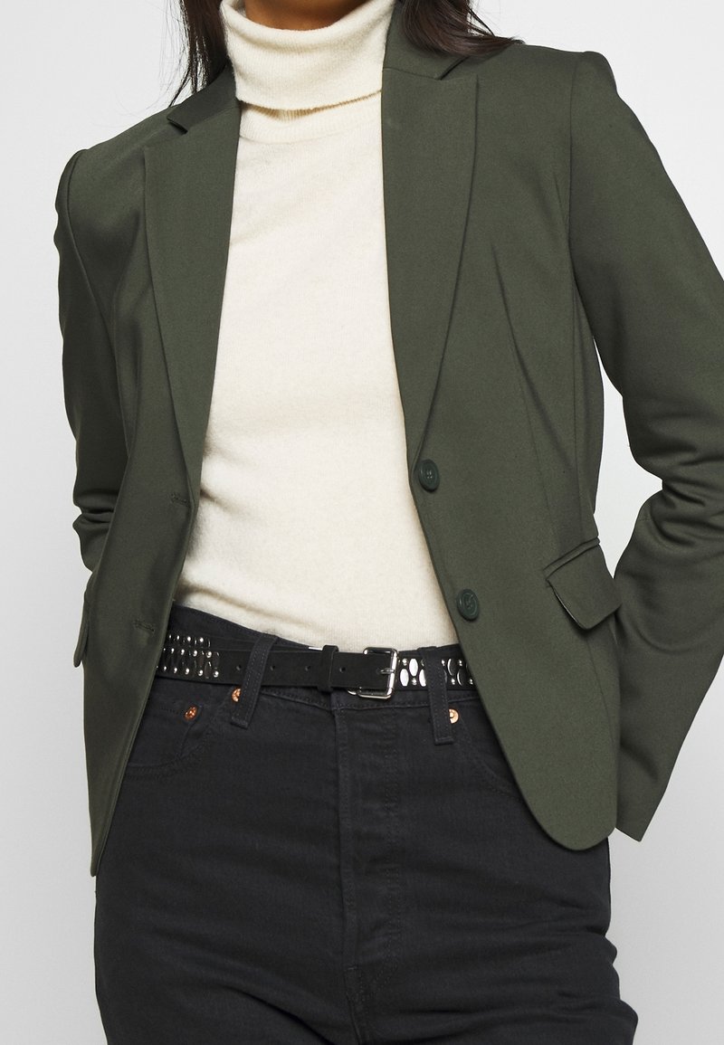 Olive green blazer with structured shoulders, two buttons, flap pockets, and a tailored fit, paired with a white sweater and black jeans.
