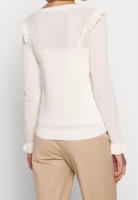 Žena v bílém pleteném svetru s dlouhými rukávy a volánovými detaily na ramenou a manžetách, oblečená s béžovými kalhotami, znázorněná zezadu.