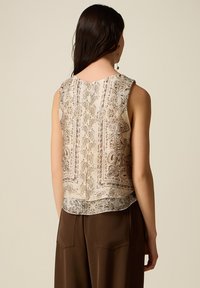 Mujer de cabello largo y oscuro, vistiendo un top sin mangas beige con estampado y pantalones marrones, vista desde atrás sobre un fondo liso.