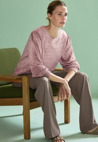 Pulover roz deschis, cu model melanj, cu decolteu în V, cu un croi lejer; asortat cu pantaloni bej, evazați. Stând pe un scaun tapițat verde.