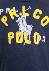 Camiseta azul marino con un gran logo bordado amarillo que dice "PRL Co POLO" acompañado de un gráfico de jugador de polo en gris contrastante.