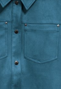 Teal mockaskjorta med knäppning fram, två bröstfickor och silverfärgade tryckknappar. Mjuk och slät textur samt design med spetstill krage.