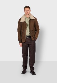 Giacca in suede marrone con colletto in pelliccia sintetica color crema, chiusura con zip e due tasche frontali, abbinata a una camicia verde chiaro e pantaloni scuri.