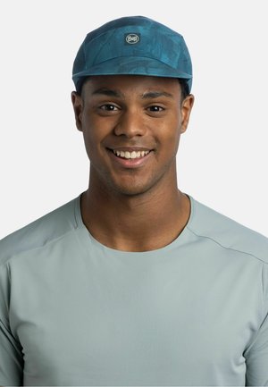 Joven sonriente que lleva una camiseta deportiva azul claro y una gorra Buff azul estampada, mirando hacia adelante sobre un fondo claro y liso.