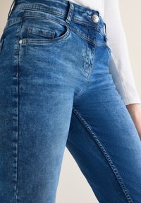 Gros plan sur un jean en denim bleu porté avec une chemise blanche à manches longues, mettant en valeur la texture et les détails de couture.