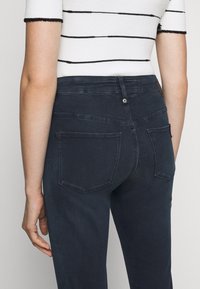 Jeans oscuros de ajuste slim, con dos bolsillos traseros, costuras sutiles y cierre de botón en la cintura. Material de algodón.