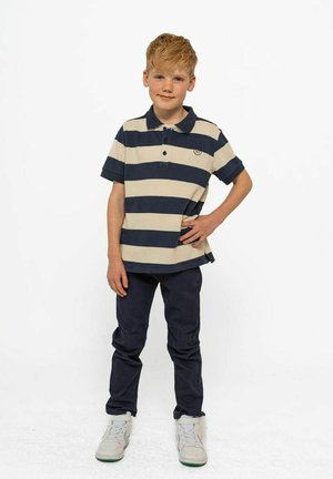 Polo à rayures beige et marine, à manches courtes, avec un col et un logo. Porté avec un pantalon bleu foncé et des baskets blanches.