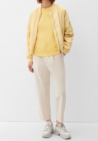 Lichtgele bomberjack met een ritssluiting aan de voorkant, geribbelde manchetten, gecombineerd met een bijpassende gele trui en beige cropped broeken. Grijze sneakers.