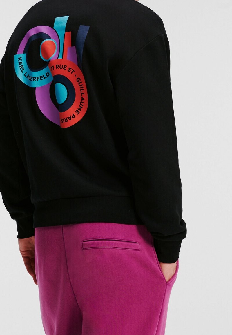 Sweat-shirt noir avec un graphique circulaire coloré et le texte "KARL LAGERFELD 27 RUE ST-GUILLAUME PARIS", porté avec un pantalon rose vif.