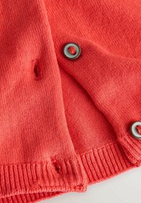Gros plan sur un tissu tricoté rouge avec deux boutons gris et deux boutonnières, un bouton partiellement attaché et une boutonnière déchirée.