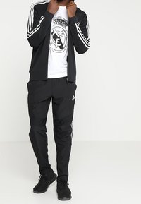 Träningskläder bestående av en svart zip-up jacka med vita ränder, i kombination med svarta byxor och en vit t-shirt med en logotyp i mitten.