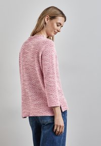 Rosa Strickpullover mit strukturiertem Finish, Dreiviertelärmeln, seitlichen Schlitzen und lockerer Passform, kombiniert mit blauen Jeans.