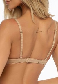 Beżowy biustonosz z teksturowanej tkaniny, regulowanymi ramiączkami i zapięciem na plecach. Posiada mały tatuaż na górnej części pleców.