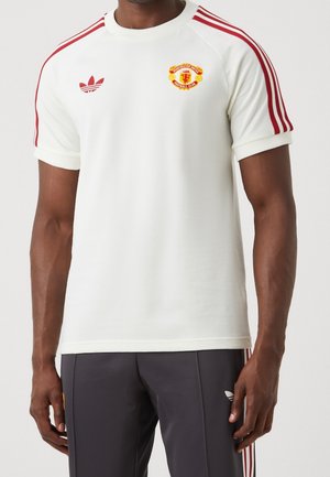 Mężczyzna ubrany w białą koszulkę Manchester United z czerwonymi paskami Adidas na ramionach oraz czarne spodnie Adidas z białymi paskami.