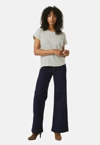 Vrouw staat in een zwart-wit gestreept shirt met korte mouwen, donkere wijde jeans en bruine enkellaarzen met hak.