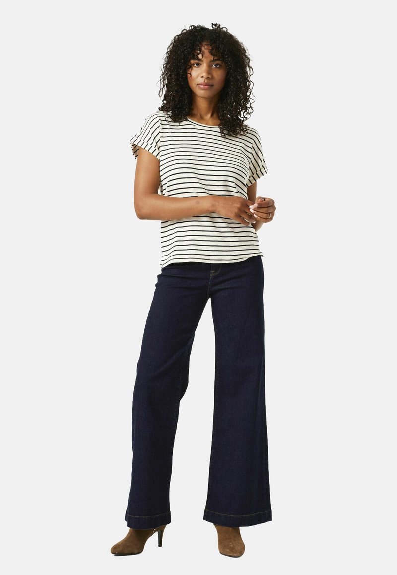 Vrouw staat in een zwart-wit gestreept shirt met korte mouwen, donkere wijde jeans en bruine enkellaarzen met hak.