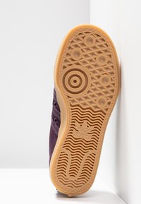 Fialová semišová teniska s texturovanou gumovou podrážkou, která má kruhové a vlnité vzory, a s vyraženým logem na spodní části podrážky.