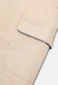 Béžový látkový rukáv s velkou klopovou kapsou, která má reflexní pruh podél okraje klopy pro zvýšení viditelnosti.