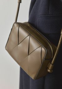 Sac bandoulière en cuir marron avec des plis géométriques, une fermeture éclair dorée et une sangle fine, présentant une texture lisse et un design structuré.
