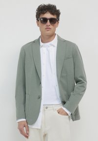 Giacca verde chiaro con texture, colletto a punta, chiusura con un solo bottone, due tasche, indossata sopra una camicia bianca e pantaloni beige. Occhiali da sole inclusi.
