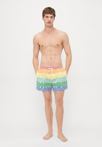 Bar överkropp man i regnbågsrandiga shorts med upprepande "DSQUARED2"-text, står barfota mot en vit bakgrund.