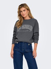Ung kvinde med langt blondt hår iført en mørkegrå sweatshirt med teksten "Los Angeles California" og højtaljede blå jeans, stående foran en hvid baggrund.