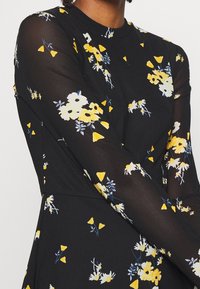 Robe noire avec manches longues transparentes ornées d'un motif floral en jaune, blanc et bleu. Encolure arrondie et silhouette ajustée.