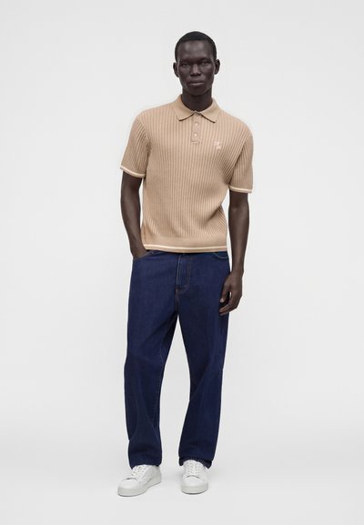 Homme portant un polo beige côtelé à manches courtes, un jean bleu foncé ample et des baskets blanches, debout devant un fond clair uni.
