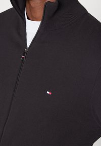 Czarny sweter zapinany na zamek z prążkowanym kołnierzem. Na piersi znajduje się małe, wielokolorowe logo, wykonany z miękkiego, teksturowanego materiału.