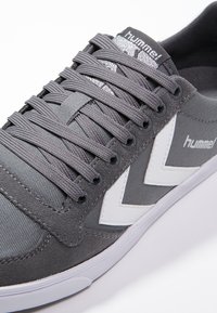 Sneaker in tela grigia con accenti bianchi a zigzag, occhielli rotondi neri e lacci testurizzati. Presenta un logo sulla linguetta.