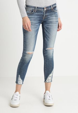 Femme portant un jean skinny déchiré bleu clair et des baskets blanches, debout devant un fond blanc uni.