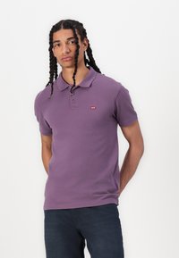 Polo din bumbac violet cu guler clasic, deschidere cu două butoane și un mic logo roșu de marcă pe partea stângă a pieptului.