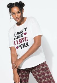 Mujer con peinado de moño espacial vistiendo una camiseta blanca con el texto "No me importa, me encanta esto" y pantalones de patrón marrón y beige.