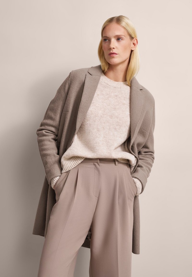 Hellgrauer Wollmantel über einem cremefarbenen Pullover und beige Farbtönen. Der Mantel hat einen Reverskragen und eine lockere Passform, mit sichtbaren Taschen.